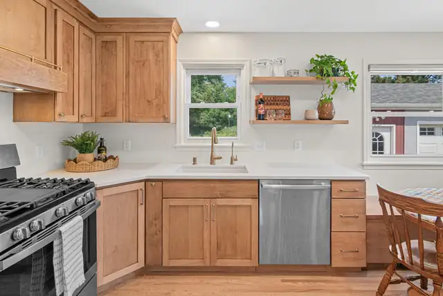 Kitchen with warm wood shaker cabinets, white counters, floating shelves and a brass faucet under the window